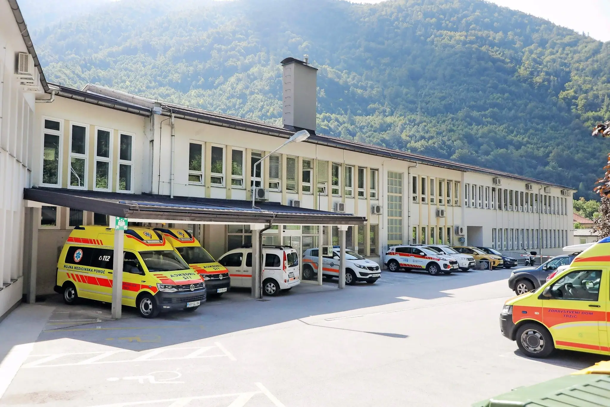 Zunanjost Zdravstvenega doma Tržič z več reševalnimi in službenimi vozili, parkiranimi pred stavbo ob vznožju gozdnate gore.